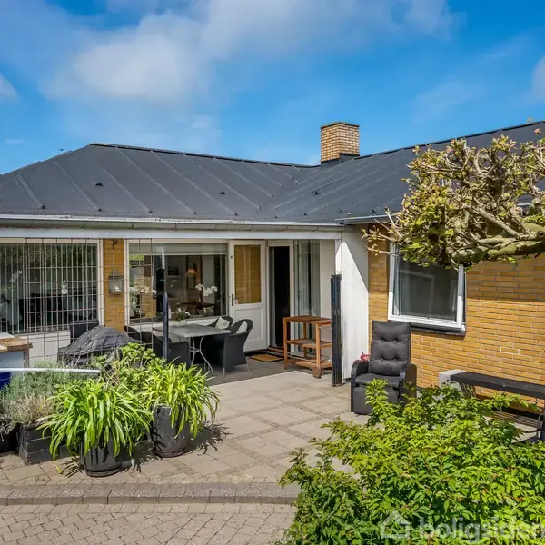Hus med fladt sort tag og gul murstensfacade, står stille i en solrig gårdhave med møbler og grønne planter. En velplejet have omkranser området under en blå himmel.