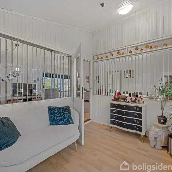 En hvid sofa med blå puder står i et rum med trægulv. Ved siden af er en kommode med dekorationer. Rummet har hvide vægge med gitre og en murstensdetalje.