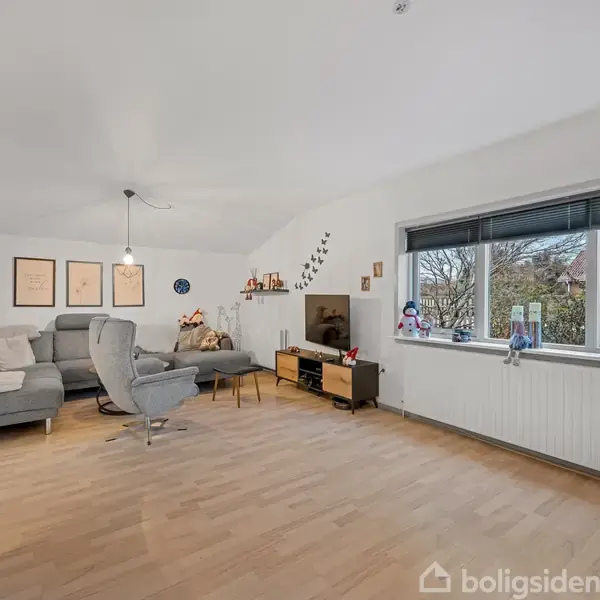 En stue med trægulv indeholder en grå hjørnesofa og en lænestol omkring et lille sofabord. Store vinduer til højre giver udsigt til en have. Væggen er dekoreret med plakater og hylder.