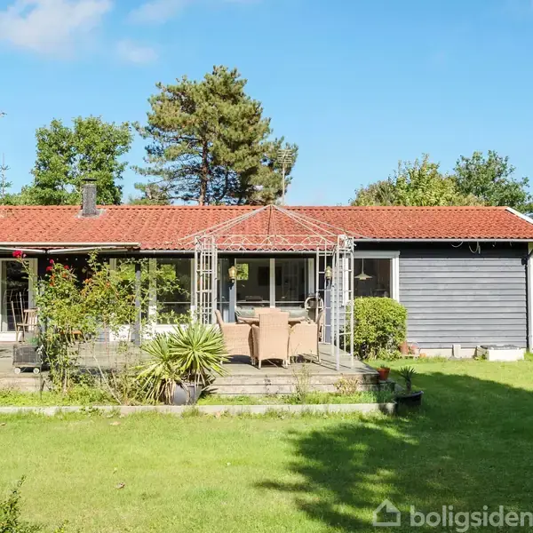 Et sommerhus med rødt tegltag og blå facade står stille i en frodig have. Foran huset ses en terrasse med havemøbler omgivet af grønne planter og træer.