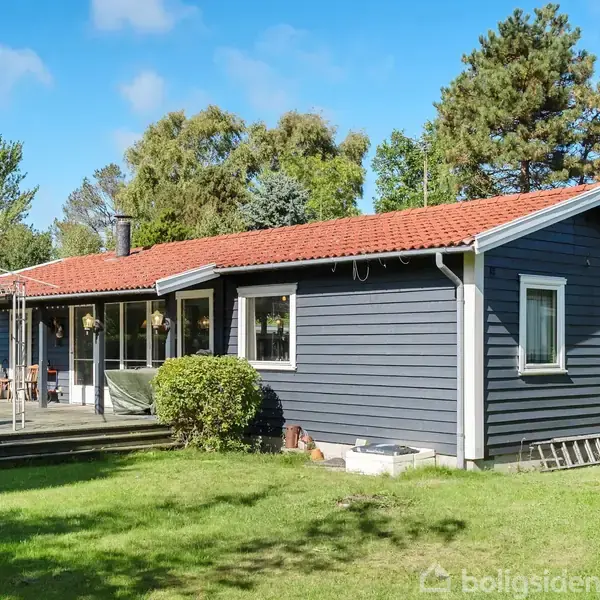Et blåt hus med rødt tag står i en solrig have. En terrasse med havemøbler ses på venstre side. Omgivet af træer og blå himmel.