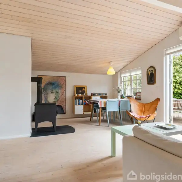 Stue med hvid sofa foran glasbord, træbeklædt loft. Spisebord og læderstol nær åbne terrasse-døre, omgivet af grønne planter udenfor. Maleri og skænk ses i baggrunden.