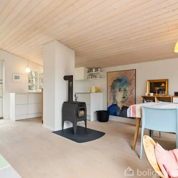 Brændeovn står midt i en moderne stue, omgivet af lyse trævægge og -loft. Bagved ses et spisebord, kunstplakat og en åben dør til køkkenet med udsigt til haven.