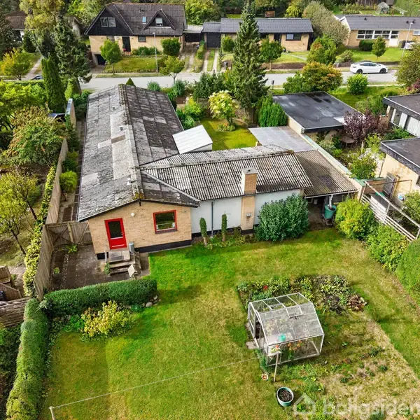 Et ældre, firkantet hus har en stor, velplejet have med buske og et fuglebur. Omkringliggende huse ses i et beboelsesområde med velholdte græsplæner og løvtræer.