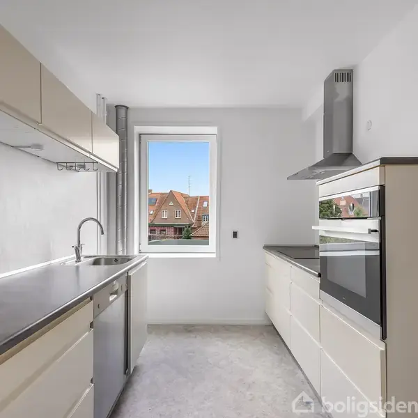 Et køkken med minimalistisk design har en vask og komfur på separate sider. Et stort vindue i midten viser gamle murstensbygninger udenfor.