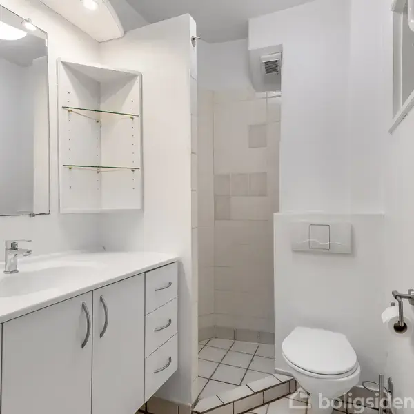 Et moderne badeværelse med hvidt skab under vasken, fliser på gulvet og toilet. Der er en bruseniche i hjørnet og et lille farverigt glasvindue på væggen.