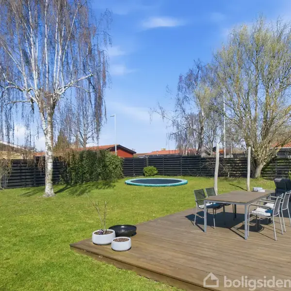 Terrassebord og stole står på en træterrasse, omgivet af en grøn have med en trampolin og store træer. Sort plankeværk omkranser området.