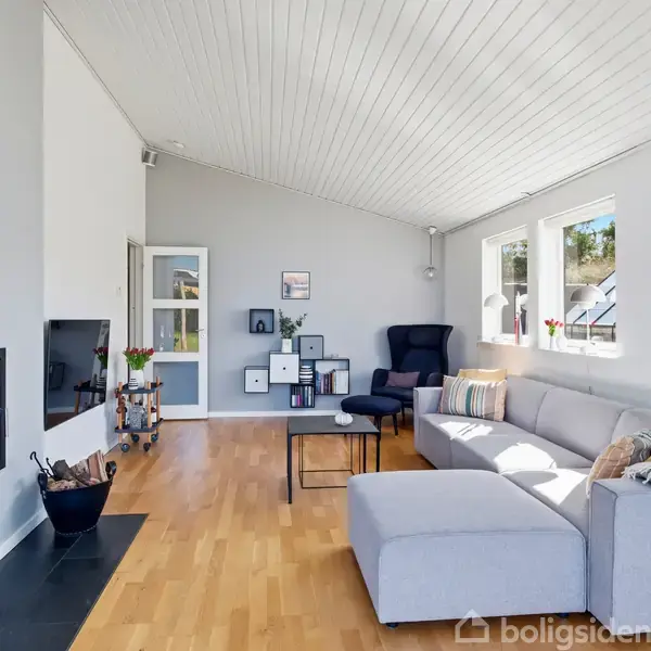 Pejs er indbygget i væg, omgivet af hyggelig stue med lysegrå sofa og sort lænestol. Store vinduer giver lys. Trægulve og minimalistisk kunst skaber stilrent rum.