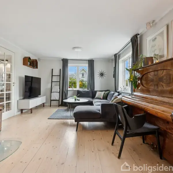 Brændeovn opvarmer en stue med lyse trægulve, hvor en hjørnesofa står ved vinduet. Et klaver står til højre, ved siden af en sort stol og dekorerede vægge.