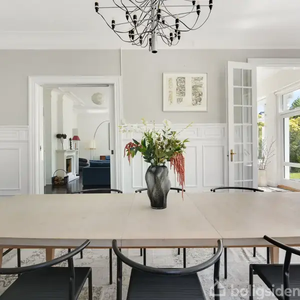 Et spisebord er dækket med en vase med blomster og omgivet af stole i et elegant rum. En moderne lysekrone hænger over bordet, og åbne døre fører til en solrig have.