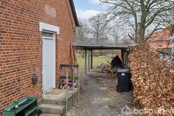 Murstensbygning med dør ses til venstre, lidt varer opbevaret ved trappen. En carport står ved siden af med en skraldespand foran. Bladløse træer og andre bygninger i baggrunden.