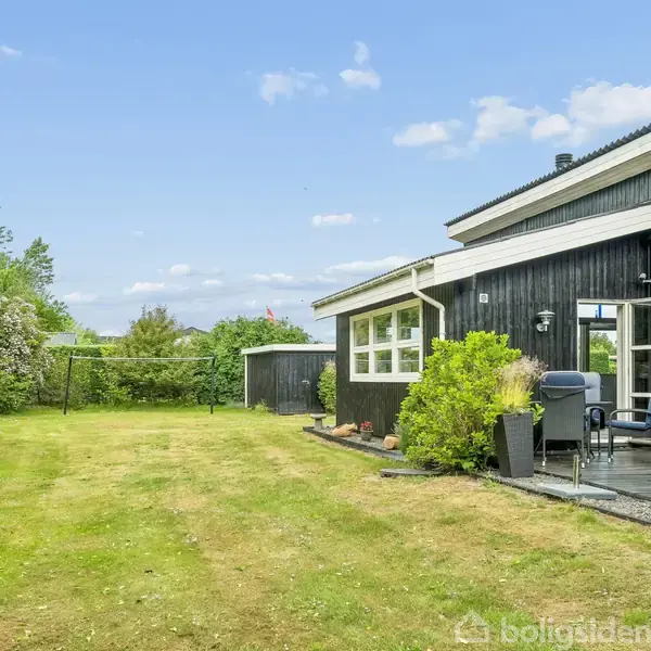 Sort hus med terrasse, omkranset af grøn have med plæne og buske. Havemøbler står på terrassen, mens himlen er blå med spredte skyer.