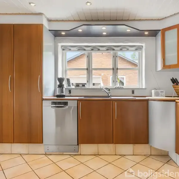 Køkken med træskabe og stål-opvaskemaskine, der står under et vindue. Rummet har lyse fliser og loftspots, mens en brødrister og knivholder står på bordet.