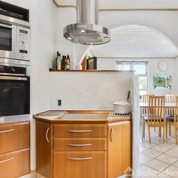 Et køkken med træskabe og indbygget ovn viser kogeplade med gryde. I baggrunden ses spisebord med stole ved et vindue. Fliser på gulvet.