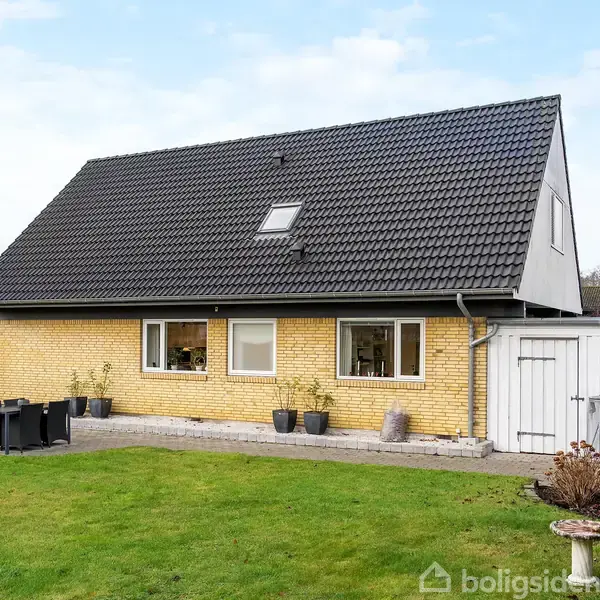 Hus med sort tegltag og gule mursten står i en velplejet have med græsplæne. Ved siden af huset er en terrasse med et sort bord og stole.