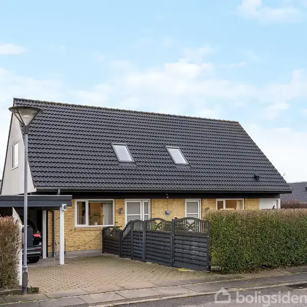Et gult murstenshus med mørkt tegltag står stille omgivet af en bøgehæk og en asfalteret indkørsel. Huset har to kviste og en carport til venstre.