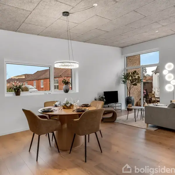 Spisebord med stole er centralt placeret i en stue med trægulv. I hjørnet står en sofa ved siden af et tv. Vinduer og planter pynter rummet.