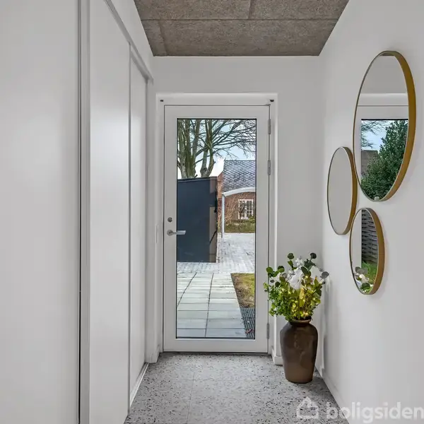 En smal gang fører til en glasdør med udsigt til en have. En vase med planter står på gulvet, og to runde spejle hænger på væggen.