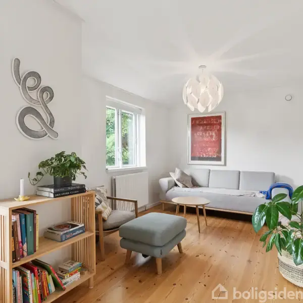 En lys stue med en grå sofa står i hjørnet ved siden af et stort vindue. En bogreol med bøger og planter er placeret på det trægulv. En dekorativ lampe hænger i midten af loftet.