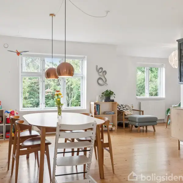 Spisebord med stole står i et lyst rum med trægulv. To hængelamper hænger over bordet. Legetøj og planter omkranser rummet, og store vinduer giver udsigt til grønne omgivelser.