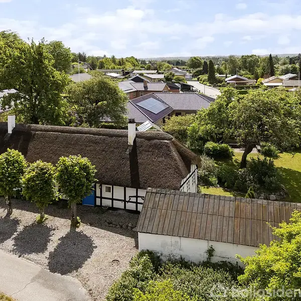 Bindingsværkshus med stråtag står stille i et grønt villakvarter med omkringliggende haver og træer. På hovedbygningens facade er der tre hvide skorstene og en mindre tilstødende bygning.
