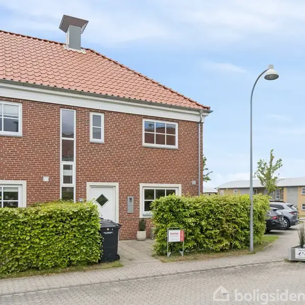 Rødt murstenshus med rødt tegltag, to etager. Hækken omkranser indgang og skraldespand. Gadebelysning og parkeret bil på villavej. Skilt ved vej giver info om boligen.
