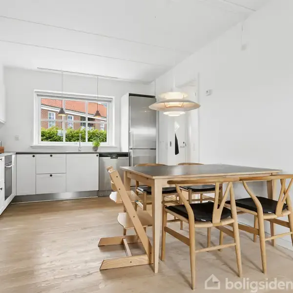 Et elegant køkken med lyse træelementer og et bord med fire stole. Et vindue giver udsigt til en bygning udenfor. Hængelampe ovenover bordet.