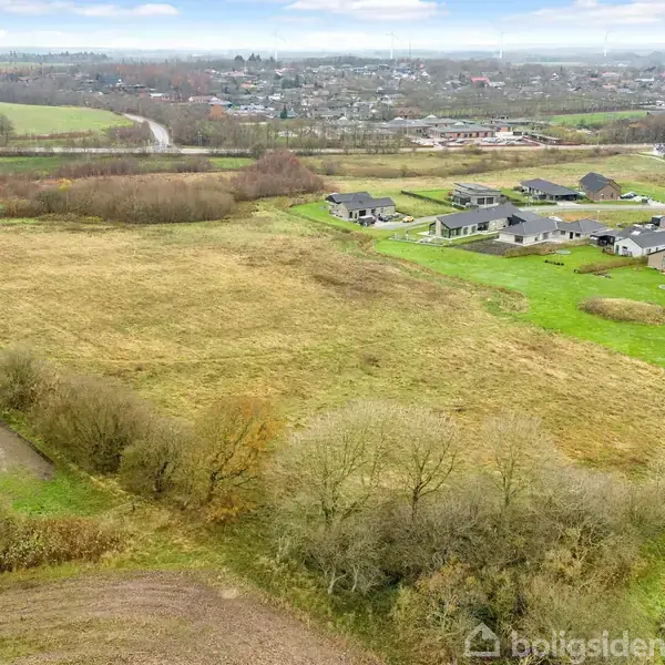 En stor åben mark strækker sig med græs og buske, omgivet af grupper af moderne huse mod højre, i udkanten af en mindre by med spredte træer og veje i baggrunden.