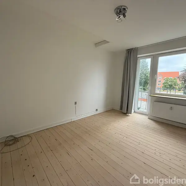 Et tomt rum med trægulv og hvide vægge. Et vindue med grå gardiner åbner ud mod en balkon, hvor bygninger og træer kan ses.