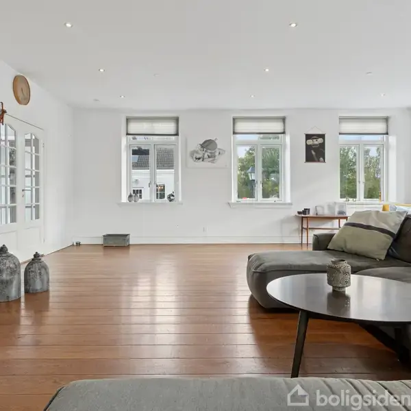 Stor stue med grå sofa, rundt sofabord og trægulv. Tre store vinduer giver naturligt lys. Vægge er hvide, dekoreret med billeder. I baggrunden står et reolsystem og pynteting.