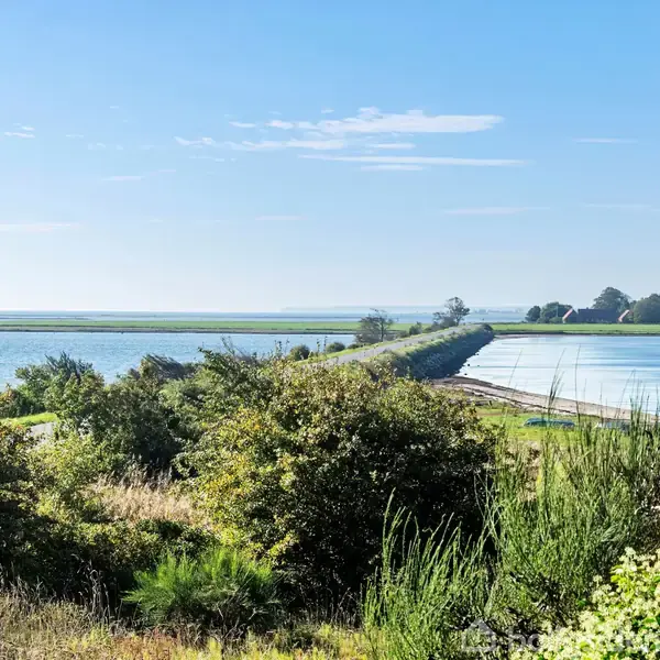 En lang vej strækker sig gennem et landligt landskab, omgivet af vand og grønt buskads, under en klar blå himmel.