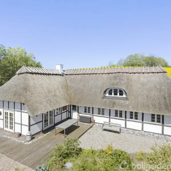 Bindingsværkshus står stille med stråtag, omgivet af blomstrende marker og grønne græsplæner under en blå himmel. Terrasse og grusgang leder rundt om huset, der har vinduer med skodder.