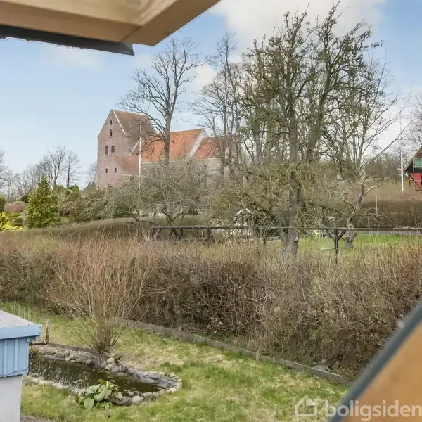 Et hus skimtes gennem en åbning i taget, omgivet af vintertræer og hække i en have. En gammel kirke med rødt tag ses i baggrunden.