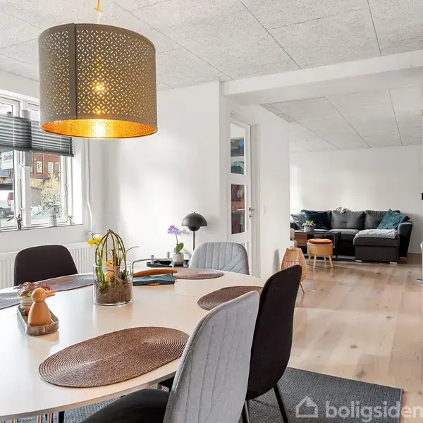 En spisestue med rundt bord er dækket med dækkeservietter og dekorationer. En stor, gul lampe hænger over. I baggrunden er en stue med sofa og reoler med glasvægge.