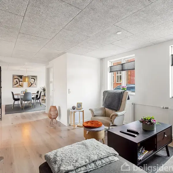 En stue forbinder til en spisestue. Sofa og lænestol står på trægulv, mens et sofabord med planter er i centrum. Store vinduer giver lysindfald.