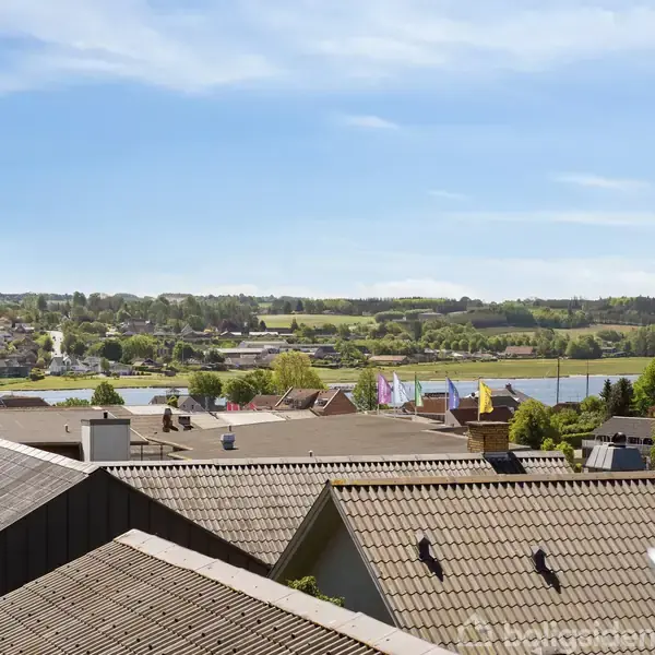 Tagtaget strækker sig over husene, der ligger tæt sammen. I baggrunden ses en flod og grønne bakker under en klar blå himmel.