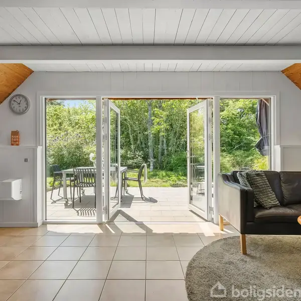 En stue med sort lædersofa og træbord står ved siden af åbne franske døre, der fører ud til en terrasse med havemøbler. Udenfor ses grønne træer og buske.
