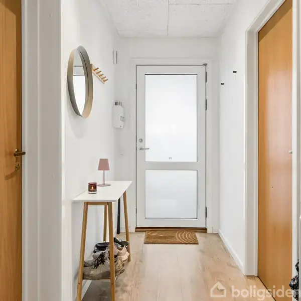 Et lyst gangområde med en hvid dør for enden, et lille bord med en lampe og spejl på væggen til venstre, og trægulv. Ingen tekst er synlig.