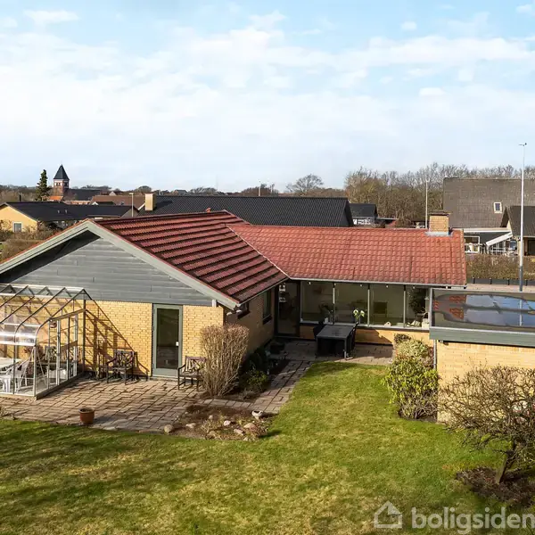 Et parcelhus med rødt tegltag har en tilhørende lukket terrasse og drivhus ved siden af. Det er omgivet af en velplejet græsplæne og ligger i et villakvarter.