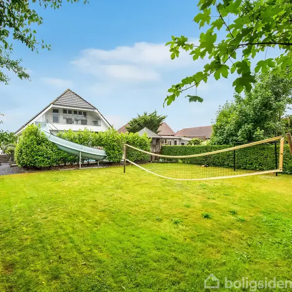 Græsplæne med volleyballnet står i en have, omkranset af buske og træer. Et hus med carport og vandrutsjebane ses i baggrunden under en blå himmel.
