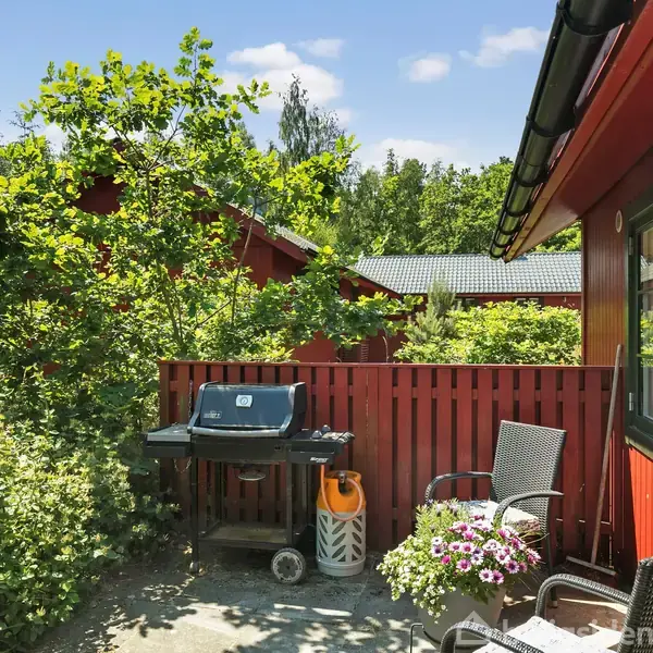 En grill står på en terrasse ved siden af en rød træbygning. Terrasseområdet har stole, blomster og er omgivet af frodig vegetation med træer og buske i baggrunden.
