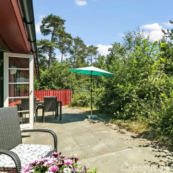 Terrassemøbler står på en flisebelagt terrasse ved et rødt hus. En parasol kaster skygge. Omgivet af grønne buske og træer, skaber det en hyggelig haveatmosfære.
