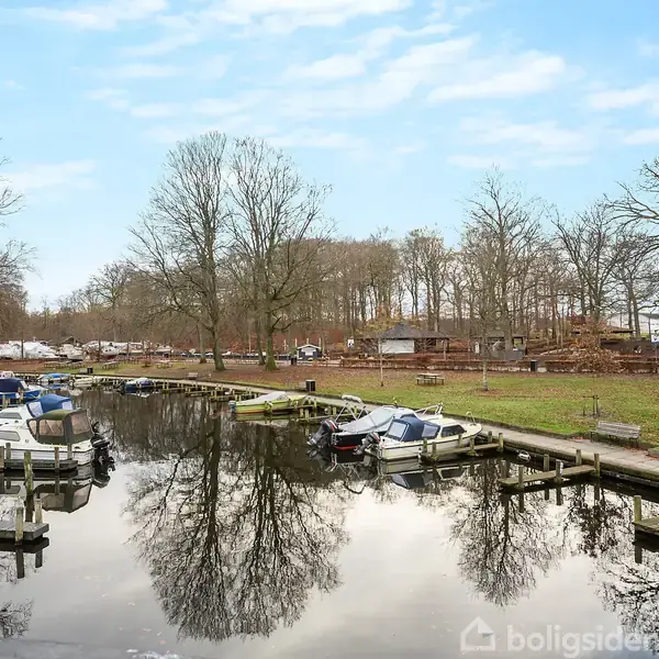 Både ligger til kaj langs en stille kanal omgivet af træer i en park. Himlen er klar, og der ses broer og græsplæner langs bredden.