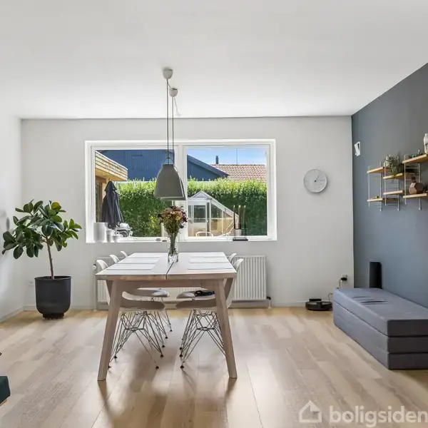 Et spisebord med fire stole står i et lyst rum. En stor plante og en billedramme er til venstre. En bænk og hylder er mod den grå væg.