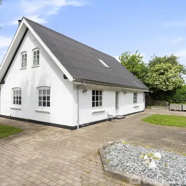 Huset er en hvidmalet bygning med sort sadeltag, omgivet af flisebelagt gårdsplads og grønne områder med træer og buske i baggrunden.