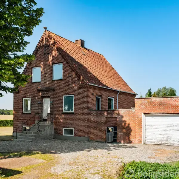 Murstenshus med rødt tegltag står på en åben grund omgivet af træer og marker. Trappe fører til hoveddør, og en garage er integreret til højre.