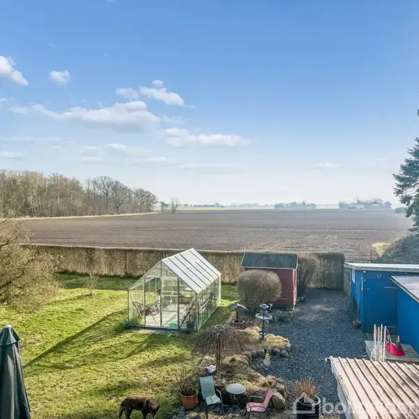 Drivhus står i en have, omgivet af græs og træer. I baggrunden ses en stor, åben mark under en blå himmel med få skyer. Der er også blå skure ved siden af det.