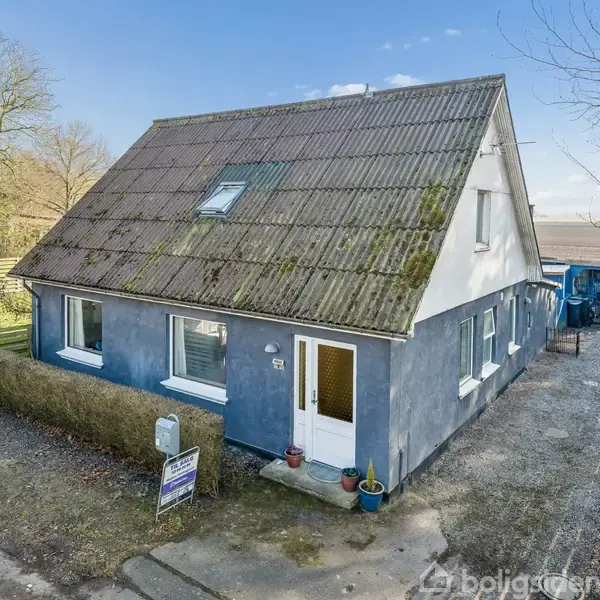 Hus med blå facade og mosdækket tegltag, står ved en landlig vej. Der er en hæk foran og indkørsel til højre. Et skilt med tekst er ved vejen.