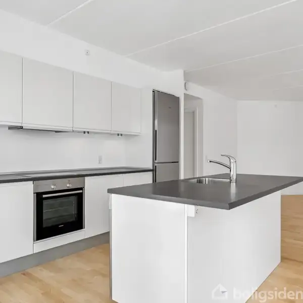 Køkkenø med vask, placeret i moderne hvidt køkken, omgivet af åbent rum med lyse trægulve og hvide vægge.