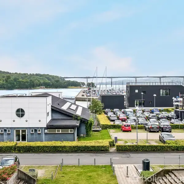 En udsigt over en by ved havet, med parkerede biler i forgrunden, moderne bygninger på hver side, og en grøn ø i baggrunden.